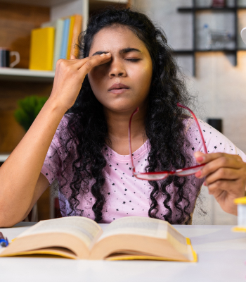 A girl holds spectacles in her left hand while touching her eyebrows with her right, with an open book in front of her.
